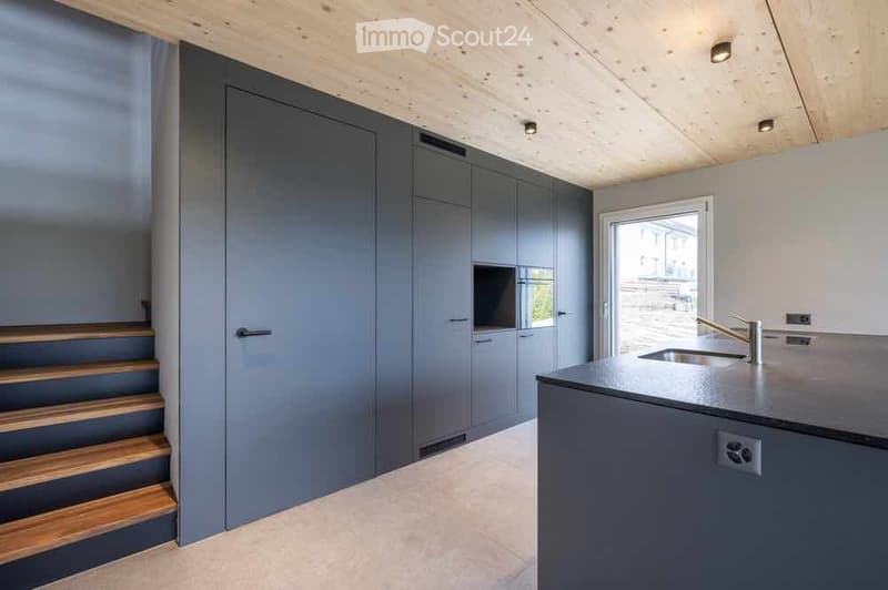 Modern kitchen with grey cabinetry, a black countertop, and a stainless steel sink. Wooden staircase leads upwards.