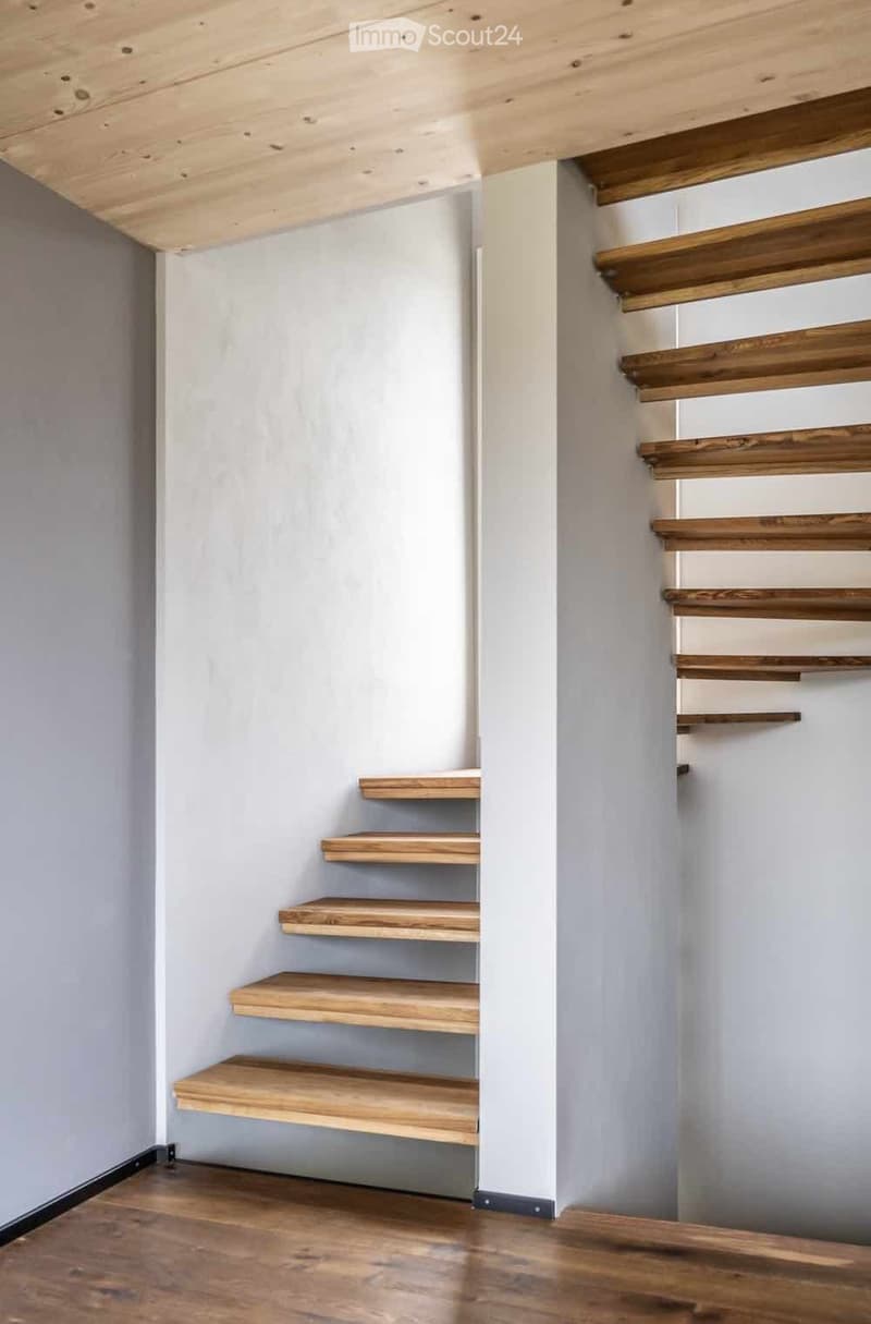 A narrow hallway with wooden stairs and built-in wooden shelves on one wall, light grey walls, and a wooden floor.