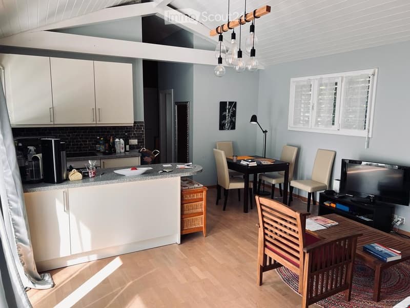 Open kitchen with breakfast bar, dining table and chairs, TV, desk with lamp, center island with appliances, wooden floors, white cabinets, white walls, windows with shutters, open beam ceiling with hanging lights.
