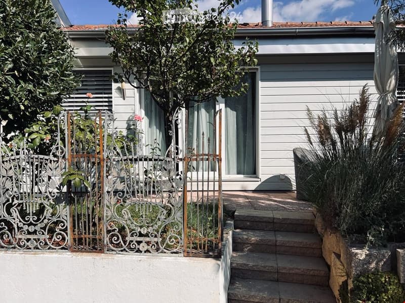 White house with red roof tiles, exterior wall with white panels, large decorative gate, concrete stairs leading to entrance, a small garden with plants and flowers