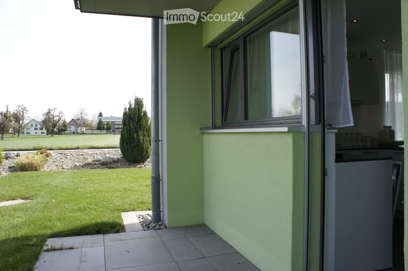 Balcony with sliding glass doors leading to a grassy yard, small garden area with rocks and shrubs, and a view of neighboring houses.