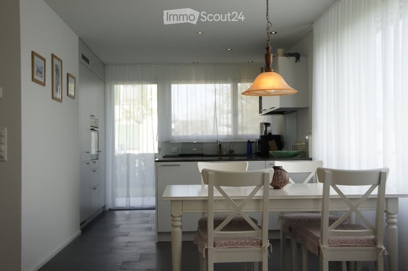 Bright dining area with a white dining table and chairs, a modern kitchen area with cabinets, appliances, and a hanging lamp.