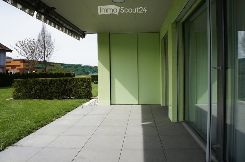 Modern balcony with large glass sliding doors, light green walls, tiled floor, and a view of a well-maintained lawn and hedge.