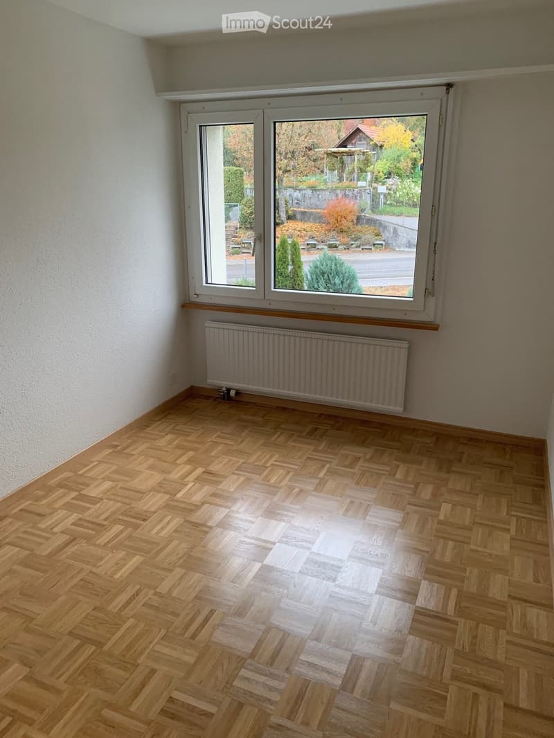 Empty room with wooden herringbone flooring, white walls, large window with view of garden, radiator below window.
