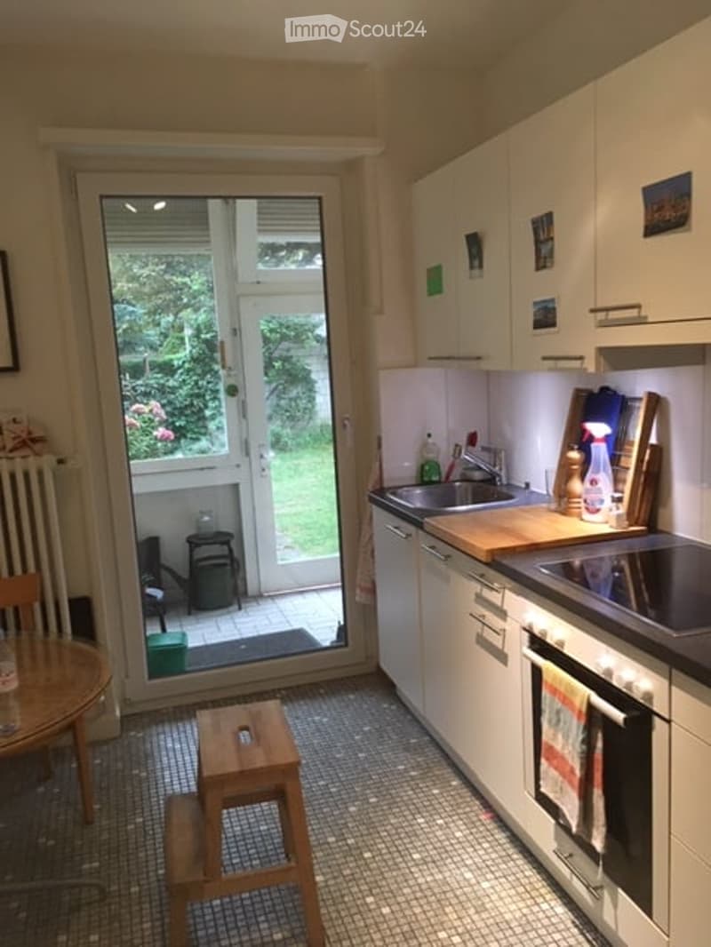 Modern kitchen with white cabinets, stainless steel sink, oven, and a small wooden stool near the door leading to a garden.