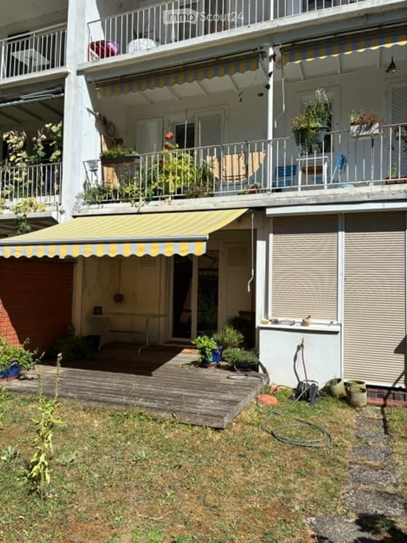 Balcony with striped awning, potted plants, and outdoor furniture, connected to a wooden deck area with additional plants and a small grassy patch.