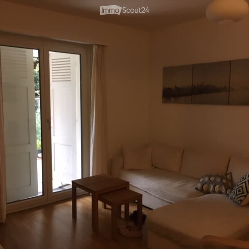 Modern living room with large windows, beige sofa, coffee table, ottoman, and artwork on the wall.