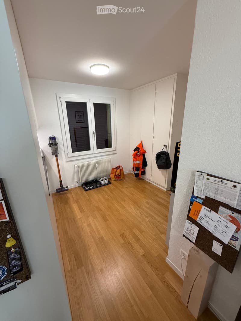 Empty hallway with wooden flooring, white walls, a window, a radiator, and a coat rack with jackets and a backpack.