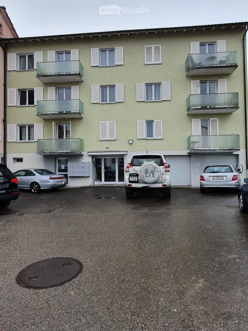 A three-story residential building with light green walls, white shutters, and multiple balconies. Several cars are parked in front of the building, and there is a manhole cover on the ground.