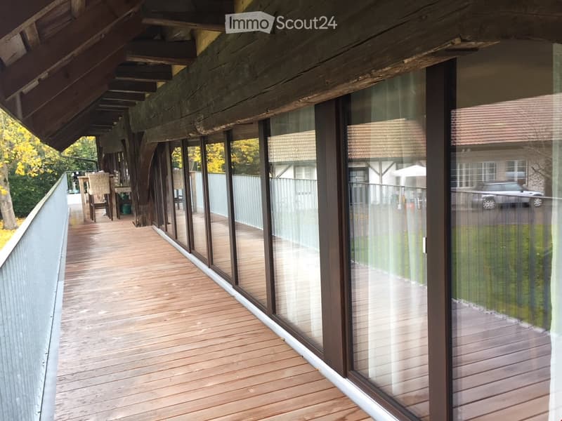 Wooden deck with glass balustrade, leading to an outdoor seating area with wicker chairs and a table.