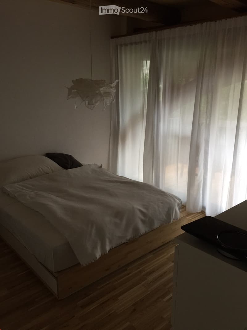 A modern, minimalist bedroom with a raised wooden bed, white bedding, and sheer curtains allowing natural light to filter in. A small table with a bowl is visible to the right.