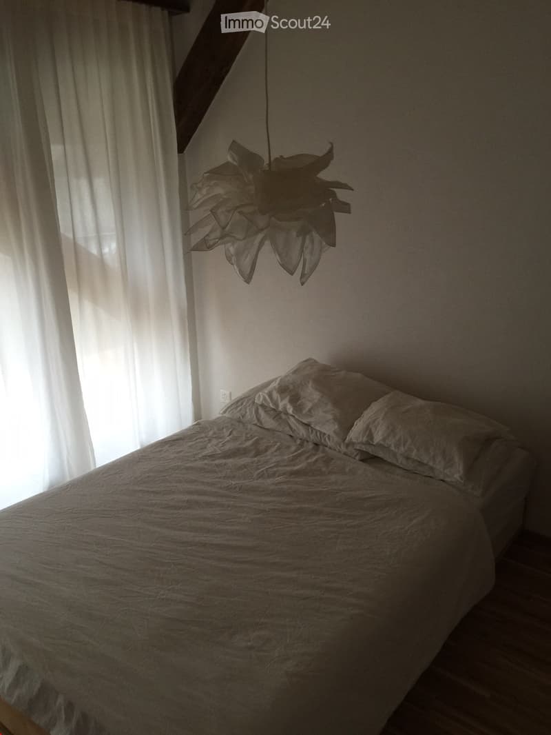 A cozy bedroom with a neatly made bed, pillows, and a hanging light fixture. The room features a large window with white curtains allowing natural light to fill the space.
