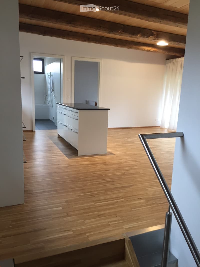 Empty room with wooden flooring, white walls, and a small kitchen counter. A staircase leads to another level, and a bathroom is visible in the background.