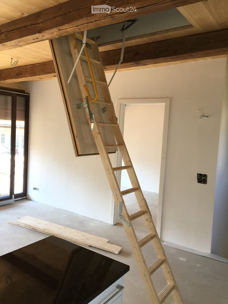 Empty room with exposed wooden beams on the ceiling, a wooden ladder leading to an attic space, and a partially visible black object on the floor.