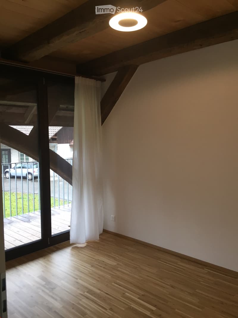 Empty room with wooden flooring, white walls, large window with white curtain, and a round ceiling light.