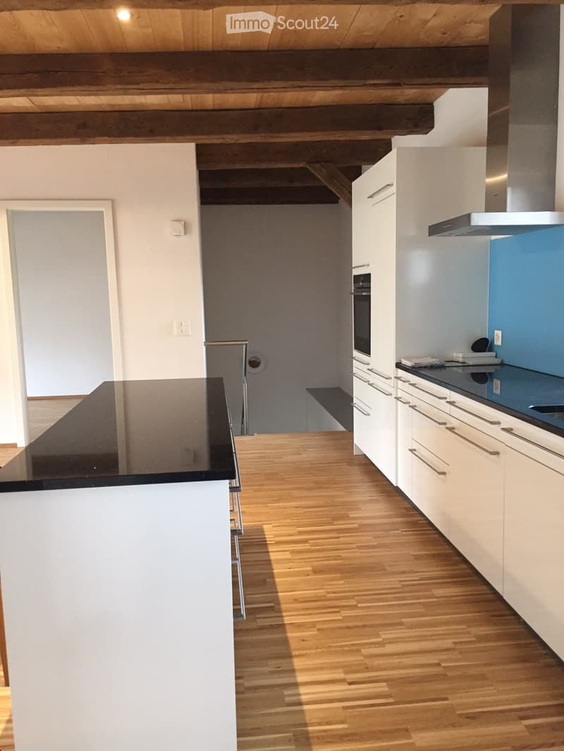 Modern kitchen with white cabinets, wooden flooring, and stainless steel appliances including a refrigerator, oven, and range hood. The countertop is black and the backsplash is blue.