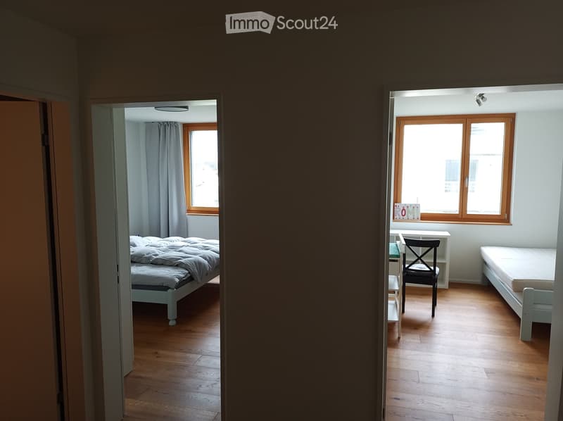Two adjacent rooms with wooden flooring, one with a bed and the other with a desk and chair, both featuring large windows with curtains.
