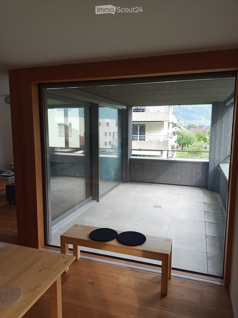 Modern living space with large glass sliding doors opening to a tiled balcony, wooden flooring, small wooden bench with two round cushions, and a small table with a stool.