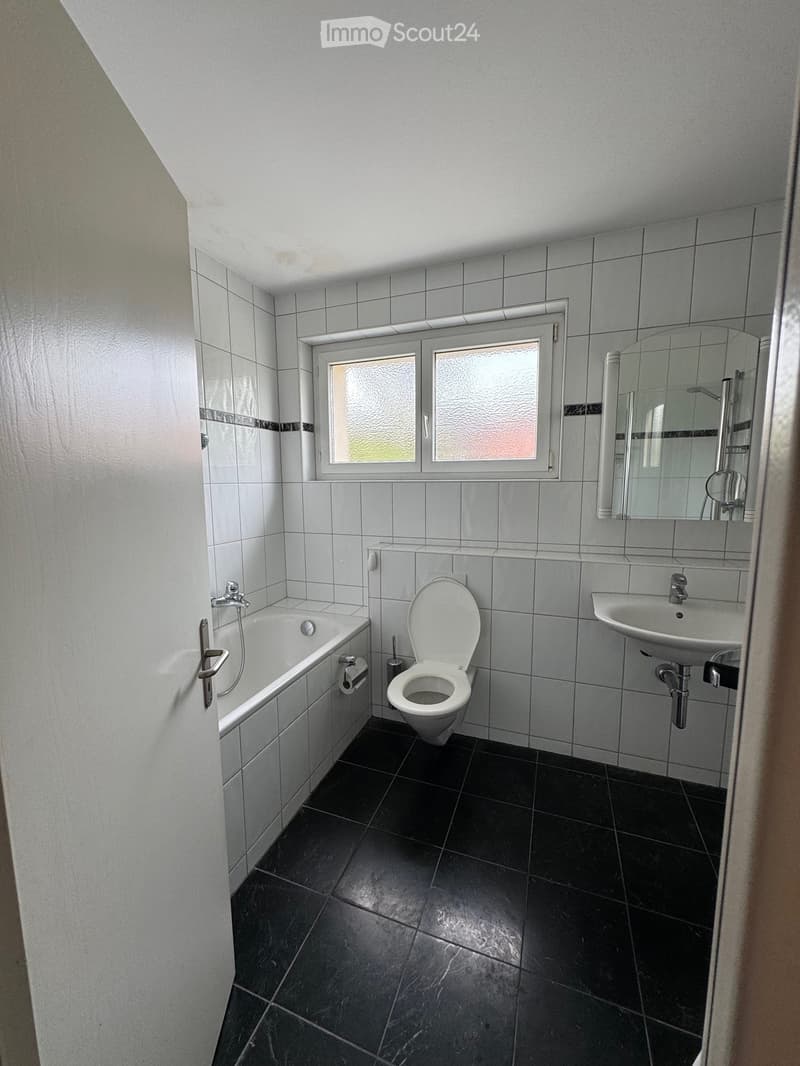 Bathroom with white tiled walls, black tiled floor, bathtub, toilet, sink, and mirror. Features a frosted window above the sink.