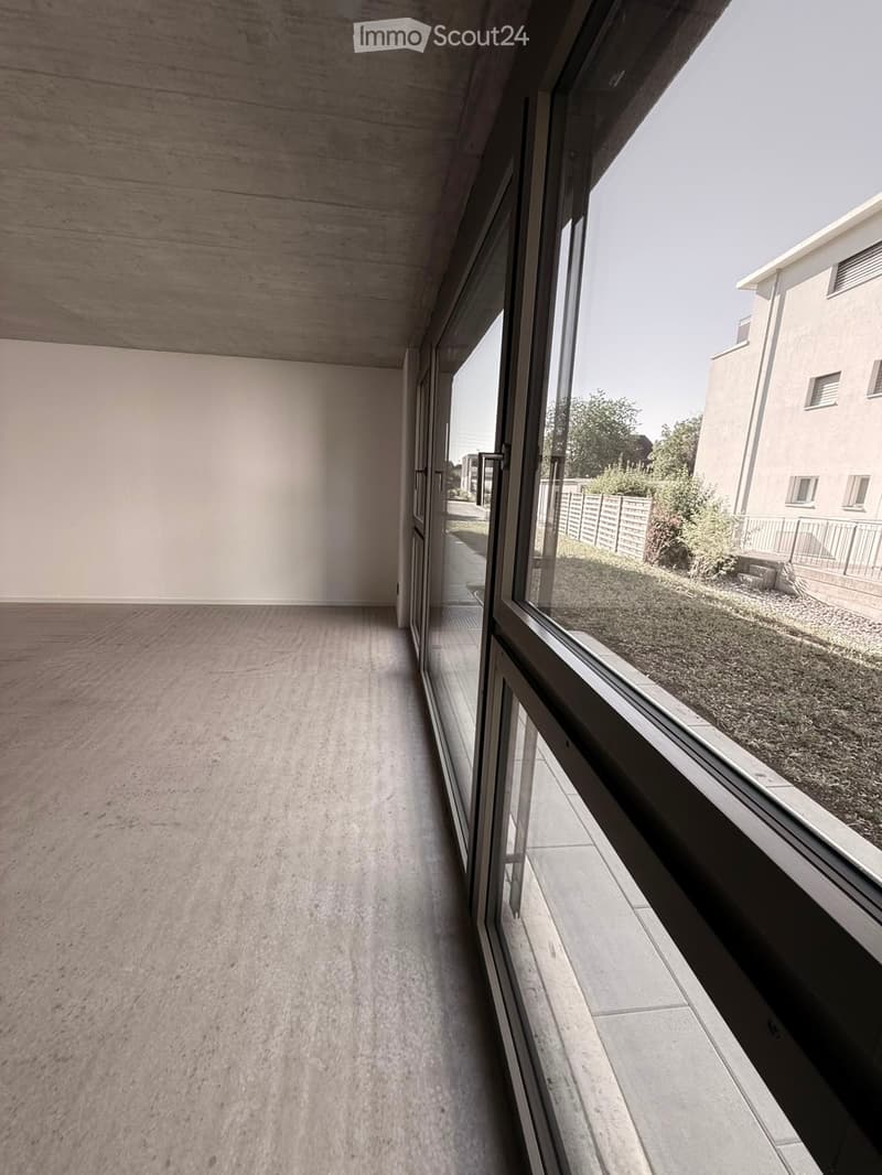 Modern, minimalist hallway with concrete floors and walls, large glass windows on one side, leading to an outdoor area with grass and a fence.