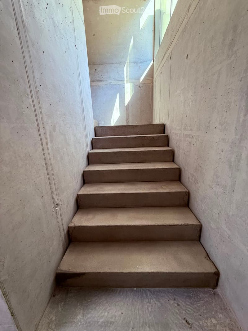 Concrete staircase with smooth steps and walls, leading upwards with natural light from a window at the top.