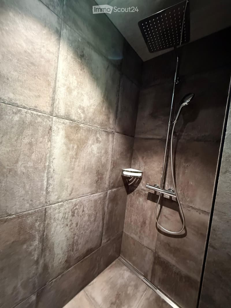 Modern shower area with grey tiled walls and floor, featuring a large showerhead and a handheld shower attachment.
