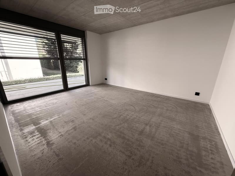 Empty room with white walls, grey carpet, large window with blinds, and visible electrical outlets.