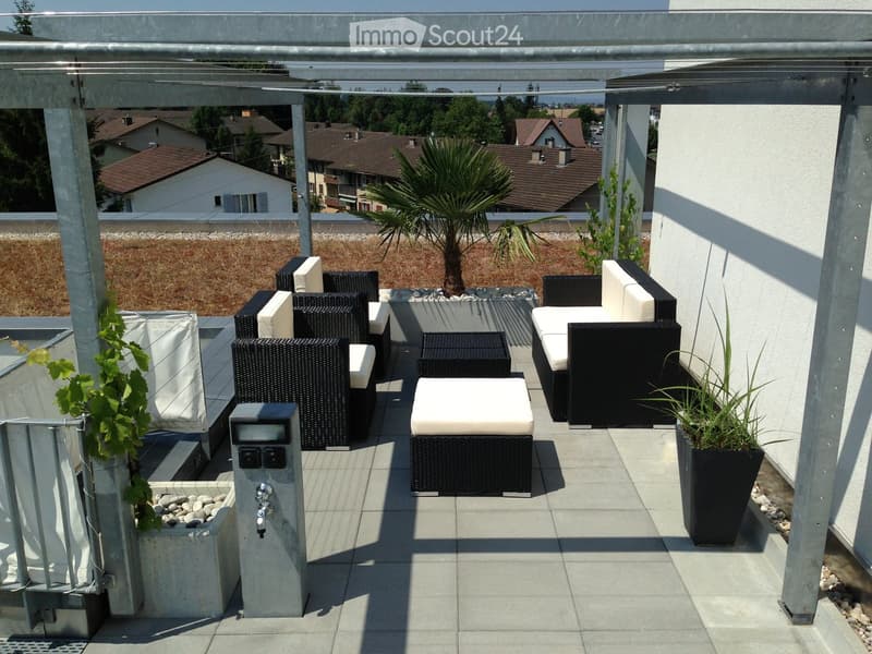 Modern rooftop terrace with wicker furniture, including a sofa, stools, and a small table, surrounded by potted plants and a view of the neighborhood.