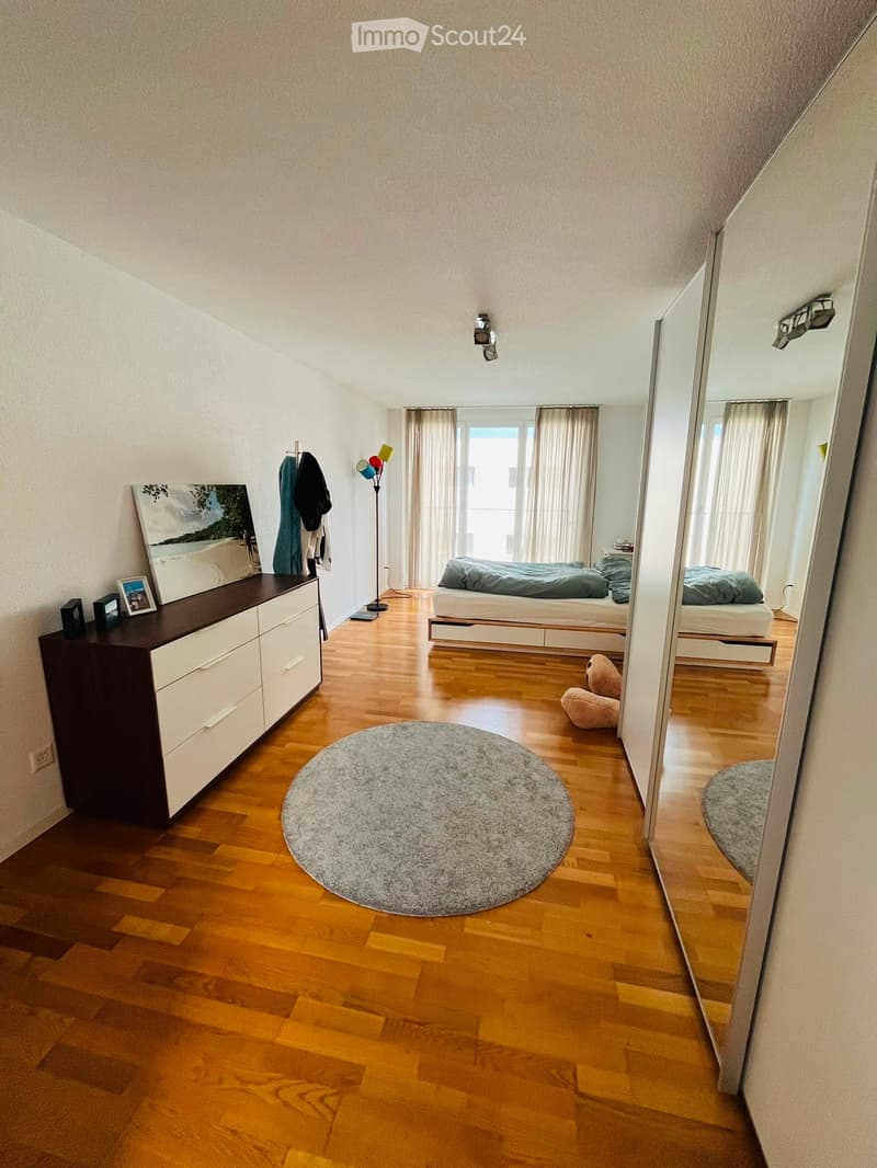 Well-lit bedroom with wooden flooring, a large bed with grey bedding, a round grey rug, a dresser with picture frames, a floor lamp, and a mirror reflecting the room.