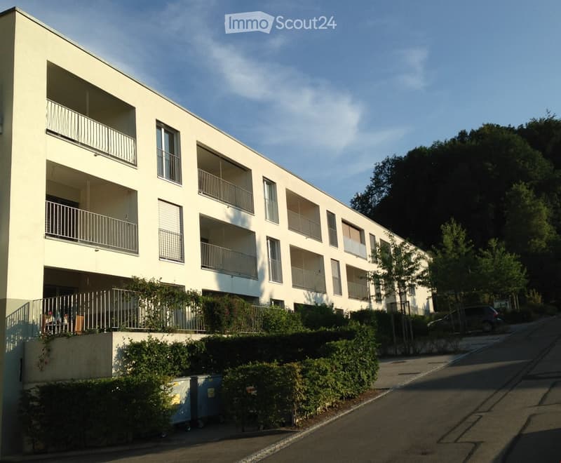 Modern multi-story residential building with balconies, large windows, and surrounding greenery.