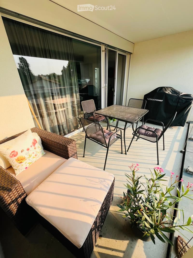 Balcony with a wicker sofa, a small table with chairs, and a covered grill. There is a potted plant with pink flowers on the floor.