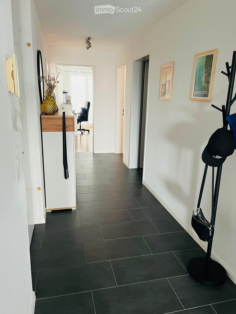 A modern hallway with dark tiled flooring, white walls, and several doors leading to different rooms. There is a coat rack with hats and bags, a small wooden desk with a vase and office chair, and framed artwork on the walls.