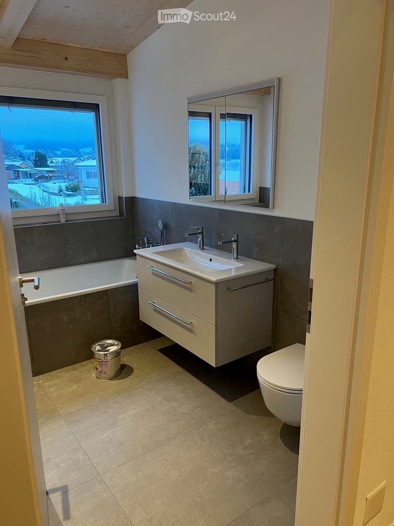 Modern bathroom with a bathtub, double sink, mirror, toilet, and cabinet. The room features large windows with a view of the outside and a slanted wooden ceiling.