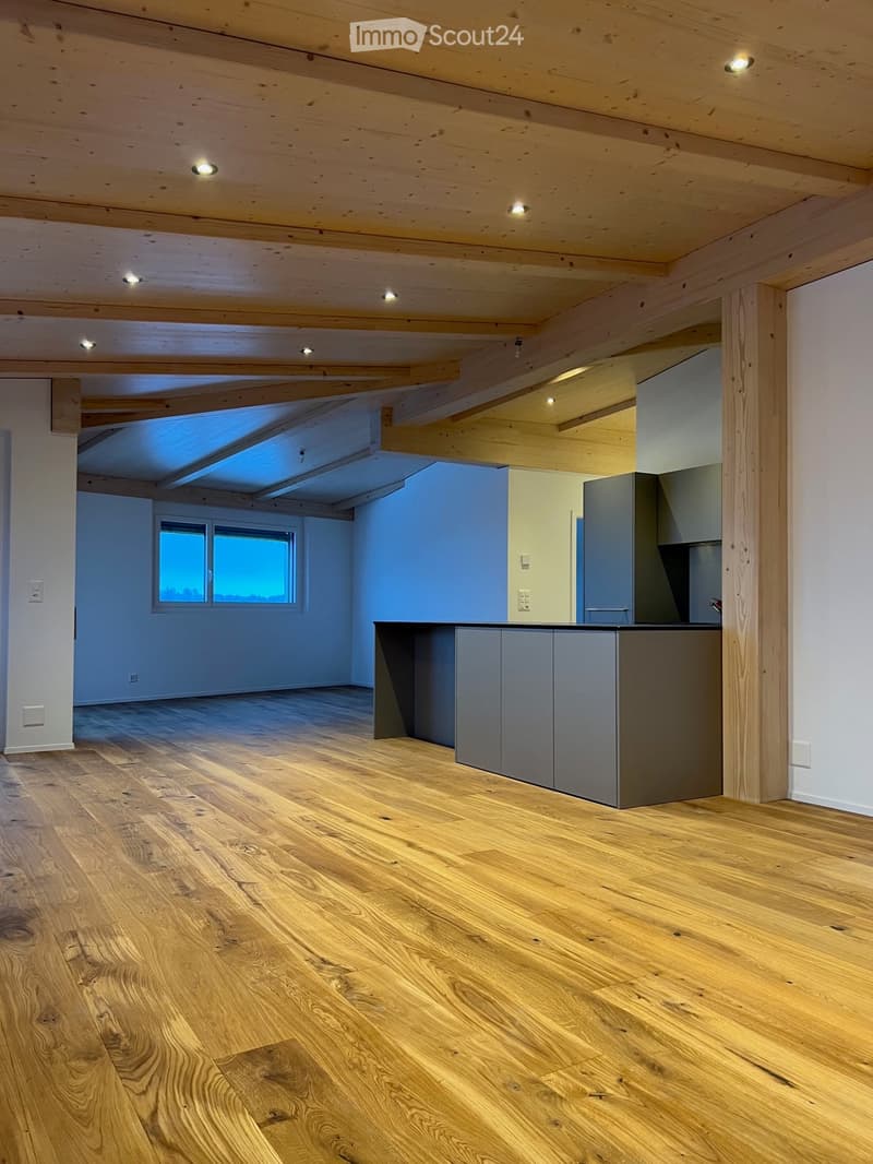 An unfurnished room with exposed wooden beams on the ceiling, wooden flooring, and a small kitchen area with cabinets and a countertop.