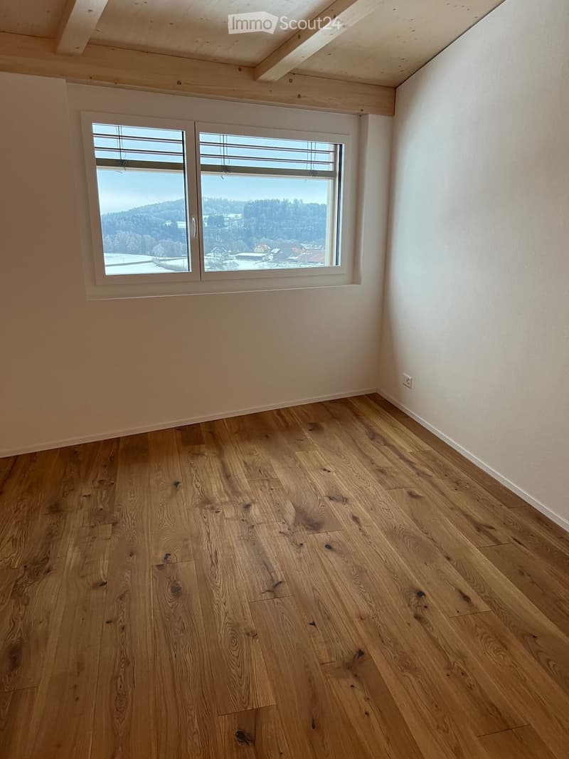 Empty room with wooden flooring, white walls, and a large window offering a scenic view of a forested area and houses.