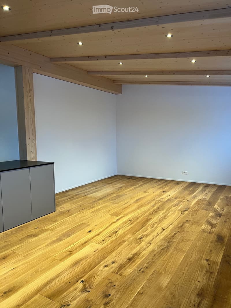 Empty room with light wooden flooring, white walls, and a small built-in cabinet on the left side. Exposed wooden beams on the ceiling with recessed lighting.