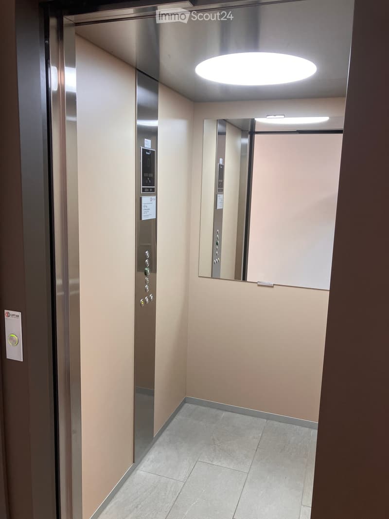 Elevator hallway with beige walls, tiled floor, and a round ceiling light. Elevator doors are closed, and control panels are visible on the walls.