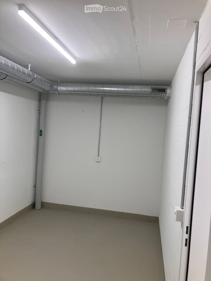Empty hallway with white walls, concrete floor, fluorescent lighting, and visible pipes and electrical outlets.