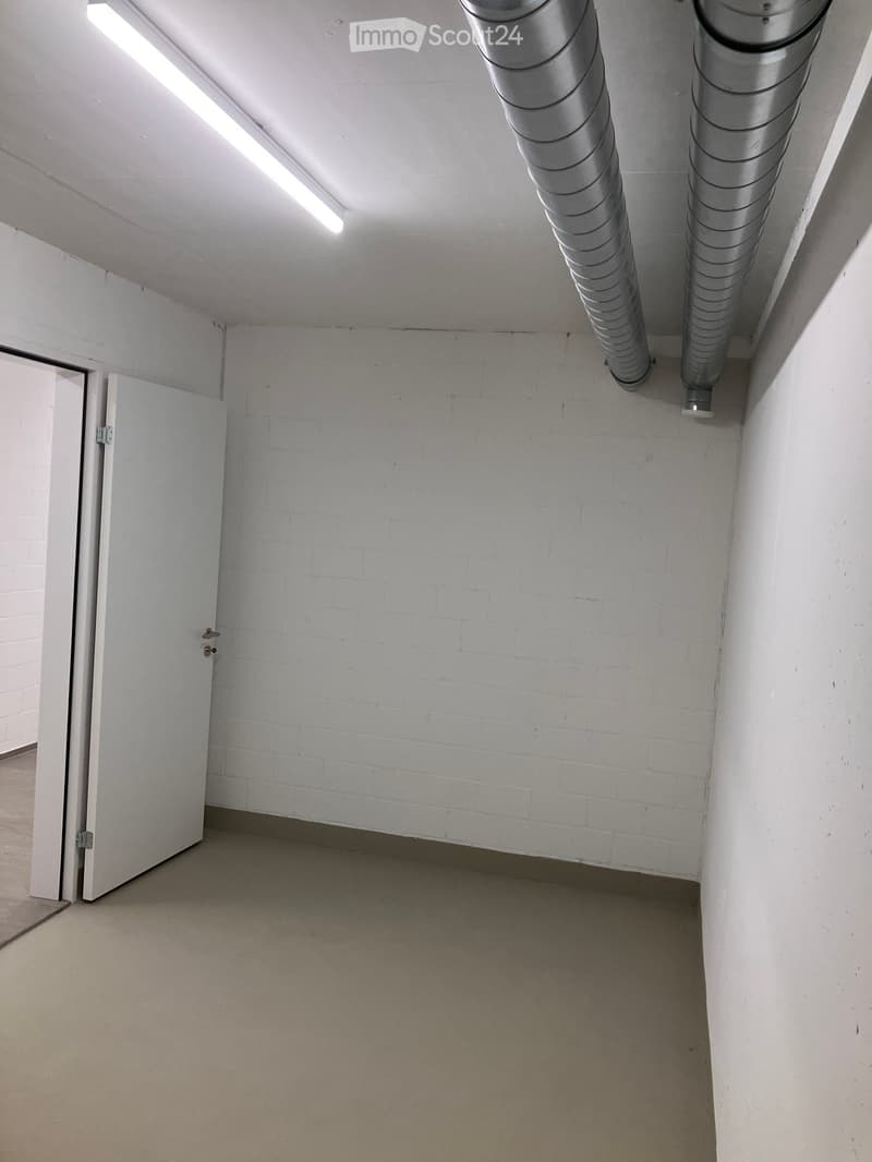 Empty room with white brick walls, concrete floor, and a single open door leading to another space. Exposed ventilation ducts are visible on the ceiling.