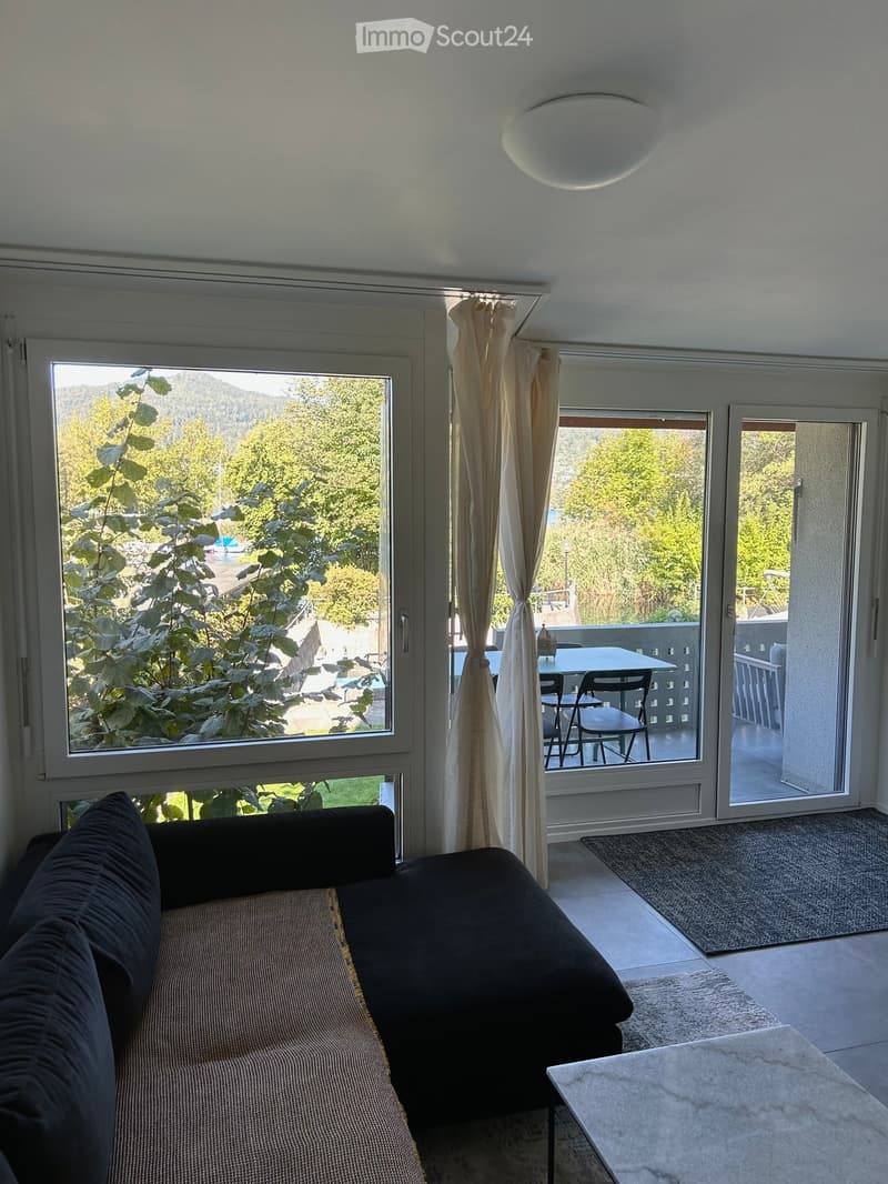 Bright living room with large windows and sliding glass doors leading to a balcony. Features a dark-colored sofa, a small table, and chairs on the balcony.