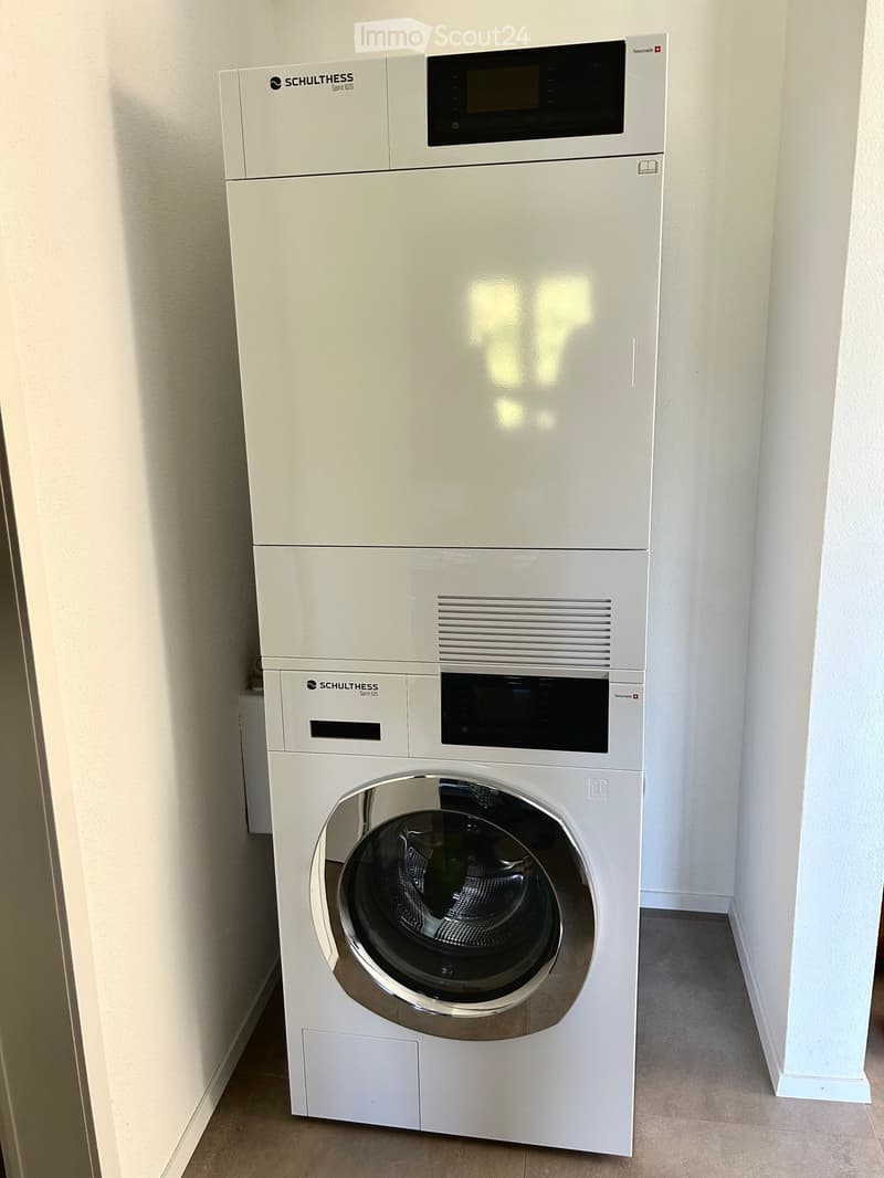 A modern, white washing machine and dishwasher unit is installed in a clean, well-lit bathroom with light-colored walls and tiled flooring.