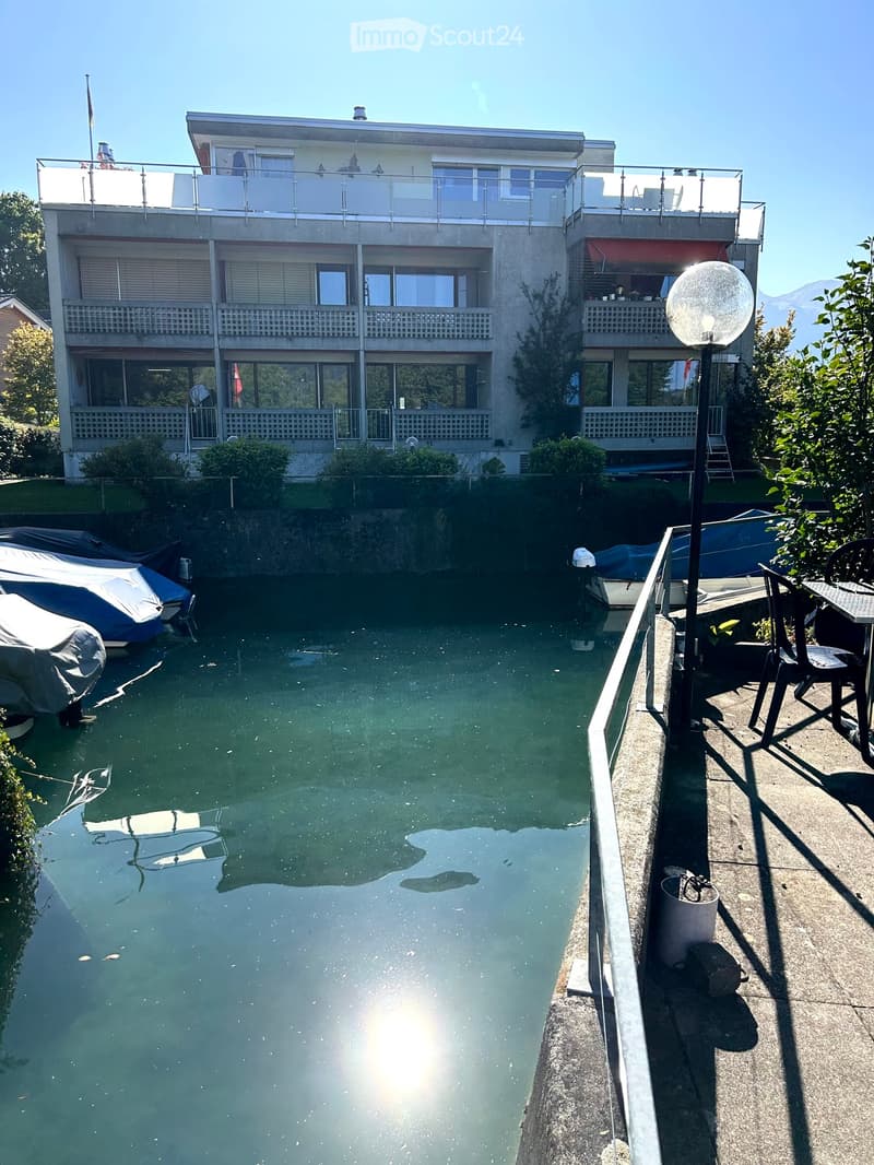 A modern multi-story house with balconies overlooking a calm canal, featuring covered boats and outdoor seating area.