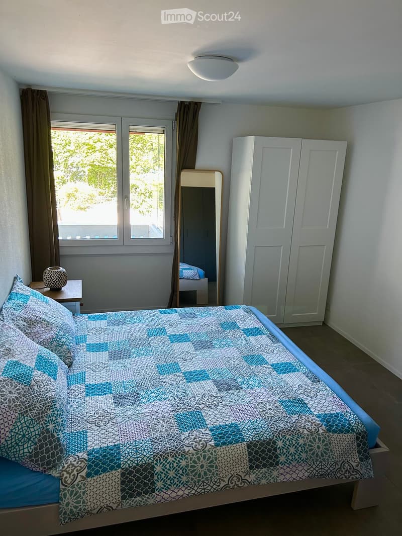 A well-lit bedroom with a double bed, nightstand, mirror, and wardrobe. The bed features a blue and white patterned duvet and matching pillows. The room has large windows with brown curtains and a wooden floor.