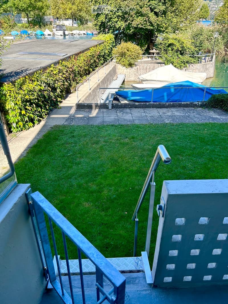 A small, well-maintained garden area with green grass, surrounded by concrete pathways and metal railings. The garden is bordered by a stone wall and overlooks a waterfront with boats and a covered seating area.
