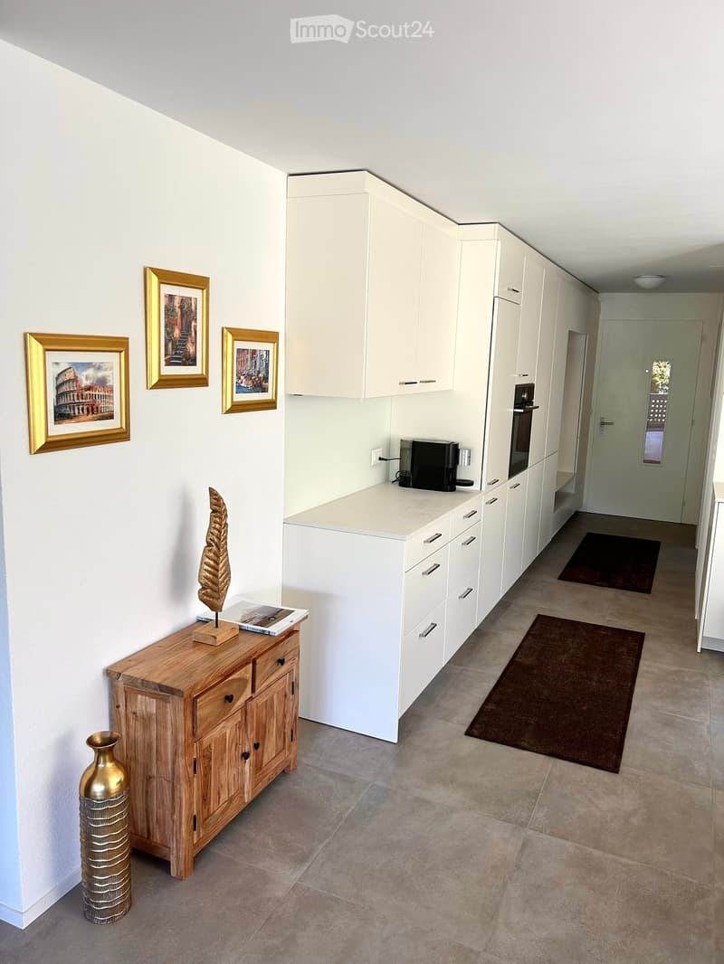 A modern hallway with a wooden cabinet, a vase, picture frames, a coffee machine, and an oven in the background.