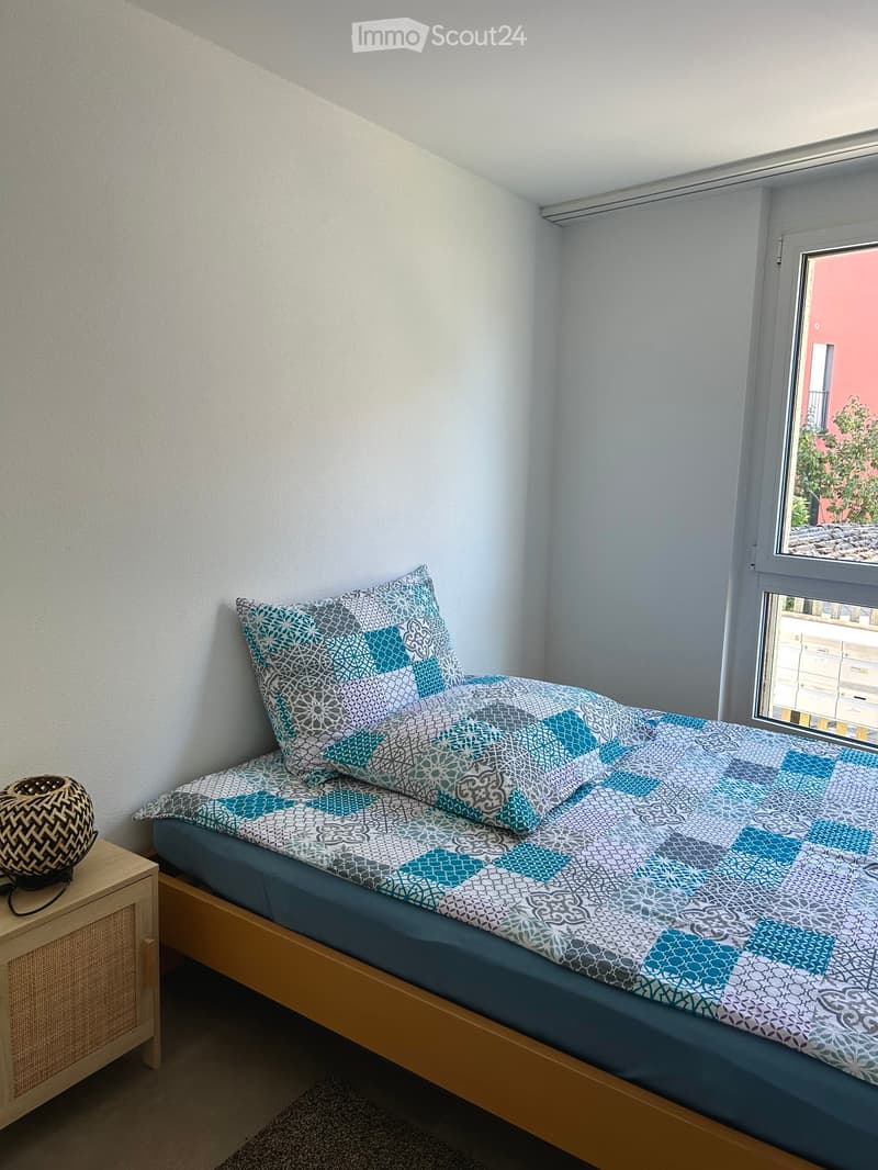 A small, cozy bedroom with a single bed, a nightstand, and a lamp. The bed is neatly made with a patterned duvet and pillows. A window with blinds allows natural light to enter.