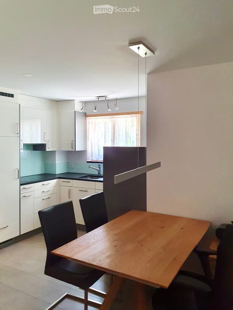 Modern kitchen with white cabinets, a wooden dining table, and black chairs. Features a sink with a faucet and a window with curtains.