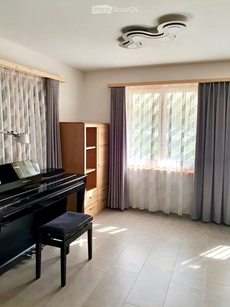 Bright room with white tiled flooring, large windows with grey curtains, a black piano with a matching stool, a wooden cabinet, and a modern ceiling light fixture.