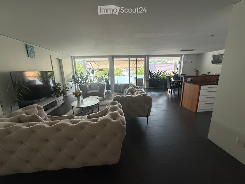 Modern living room with a large sectional sofa, coffee table, dining area, and kitchen counter. Large windows provide natural light and access to a balcony with outdoor seating.