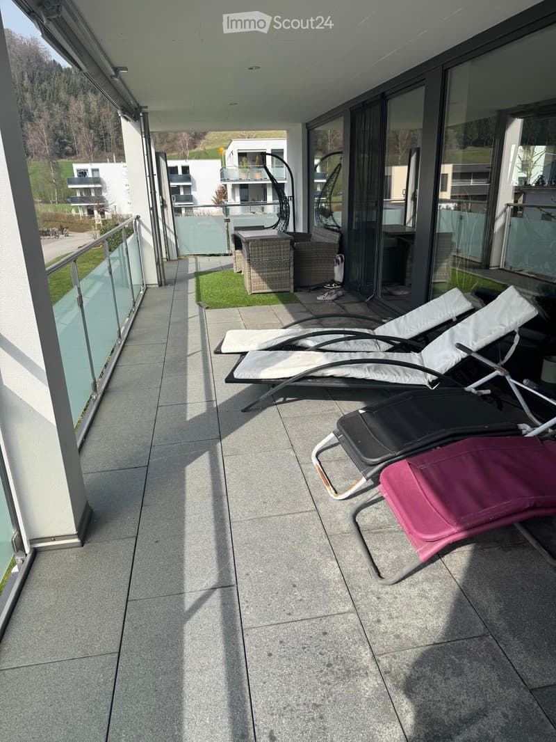 Balcony with tiled floor, glass railing, sun loungers, wicker chair, and small table.